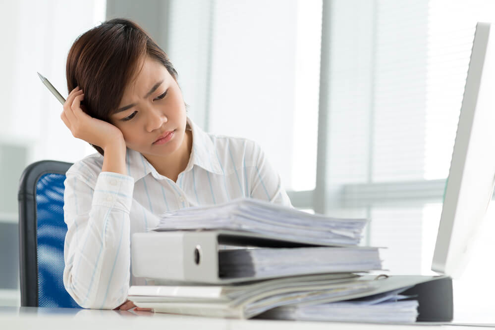 picture of a pile of binders and books and a woman with her head in hand looking stressed